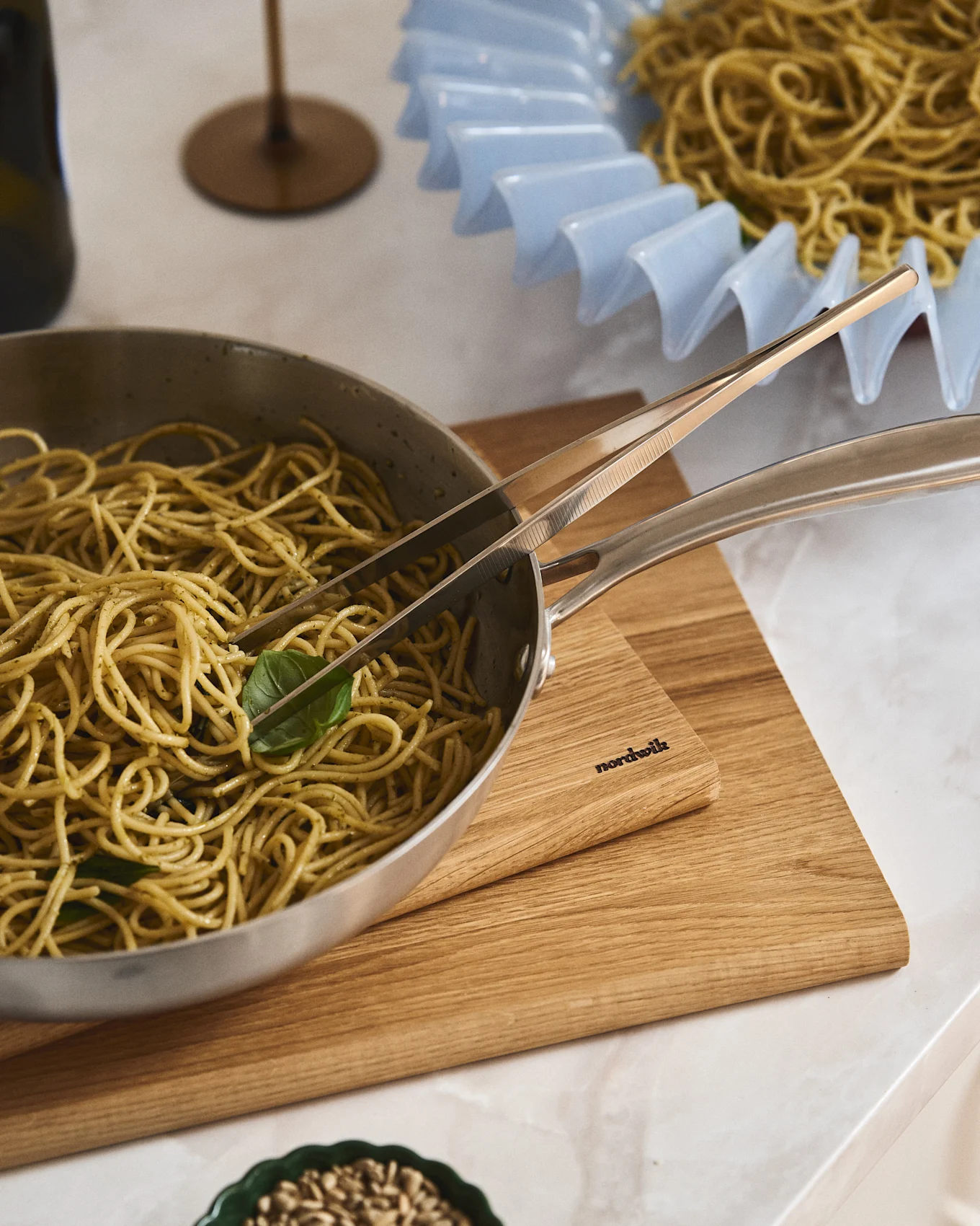 Pesto spaghetti in a silver pan with tongs on a Nordwik wooden board. Another plate of spaghetti in a light blue ruffled bowl.