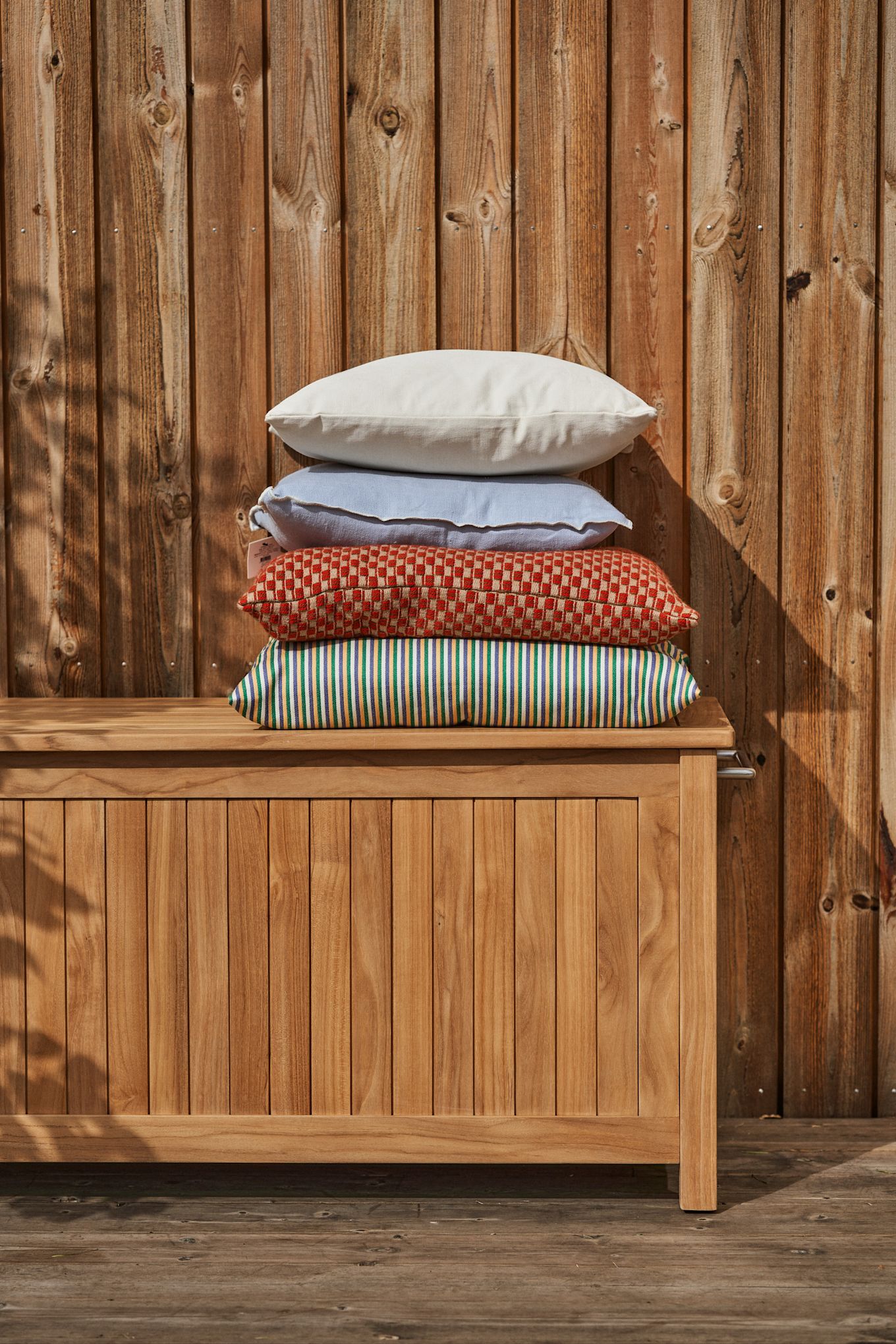 Add life, colour and comfort to your patio with cushions in mixed colours, patterns and materials, here cushions in beige, light blue, red and green on top of a teak cushion box.