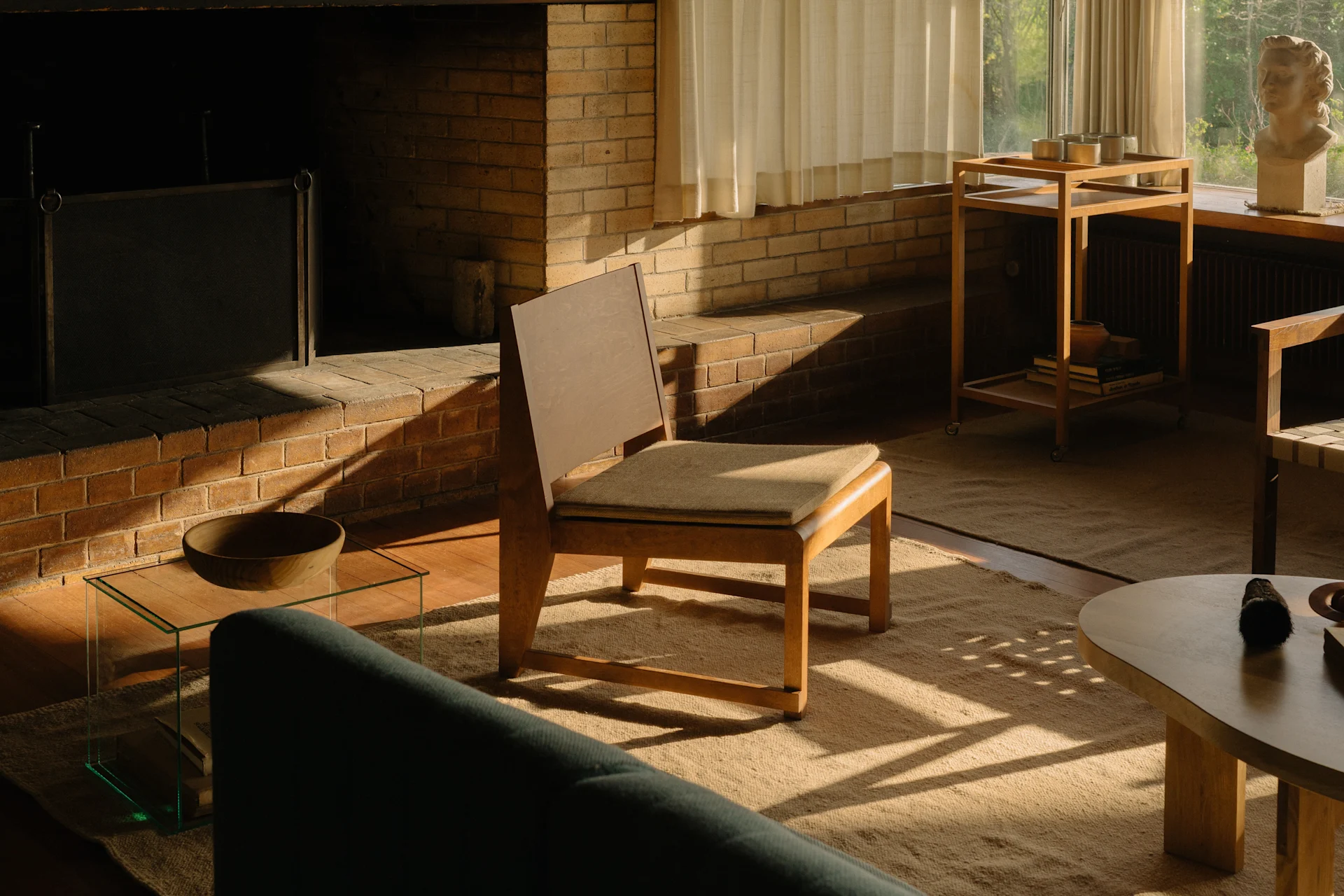 A sunlit living room with a brick fireplace, a wooden chair, and a green sofa, featuring a bust sculpture by the window.