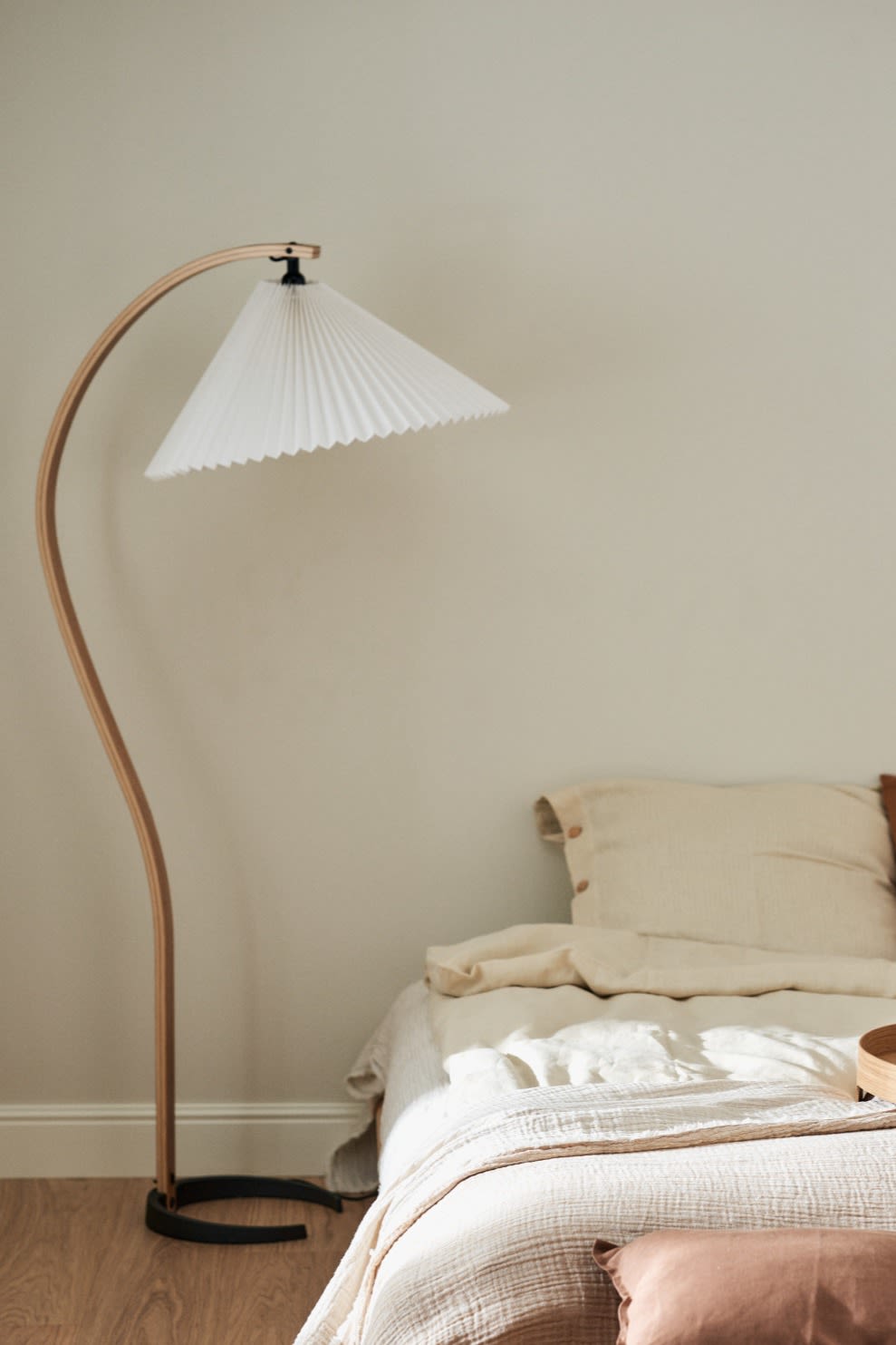 Refresh your home with these modern lighting ideas. Here you see the Gubi Timberline floor lamp with its curved wooden leg, standing next to a bed. 