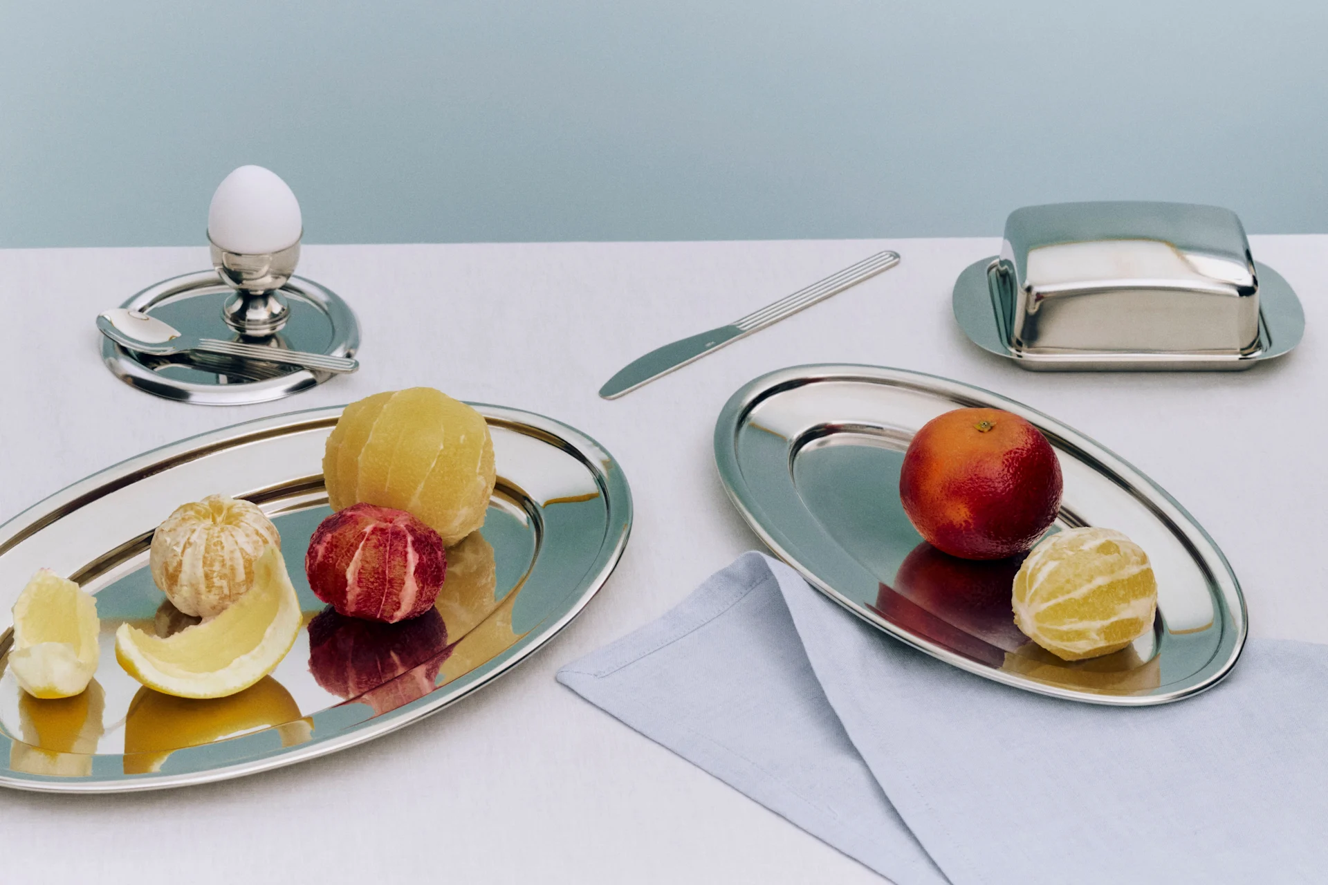 Breakfast setting with various peeled and unpeeled citrus fruits on silver trays, an egg in an egg cup, and a butter dish.