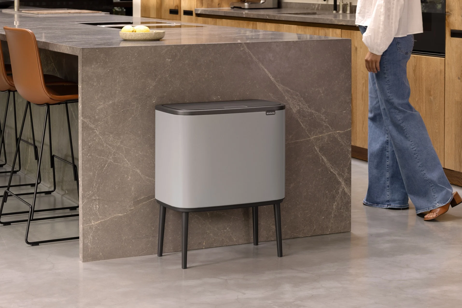 A grey bin with a dark lid on legs stands in a modern kitchen next to a brown marble island. A person walks by.