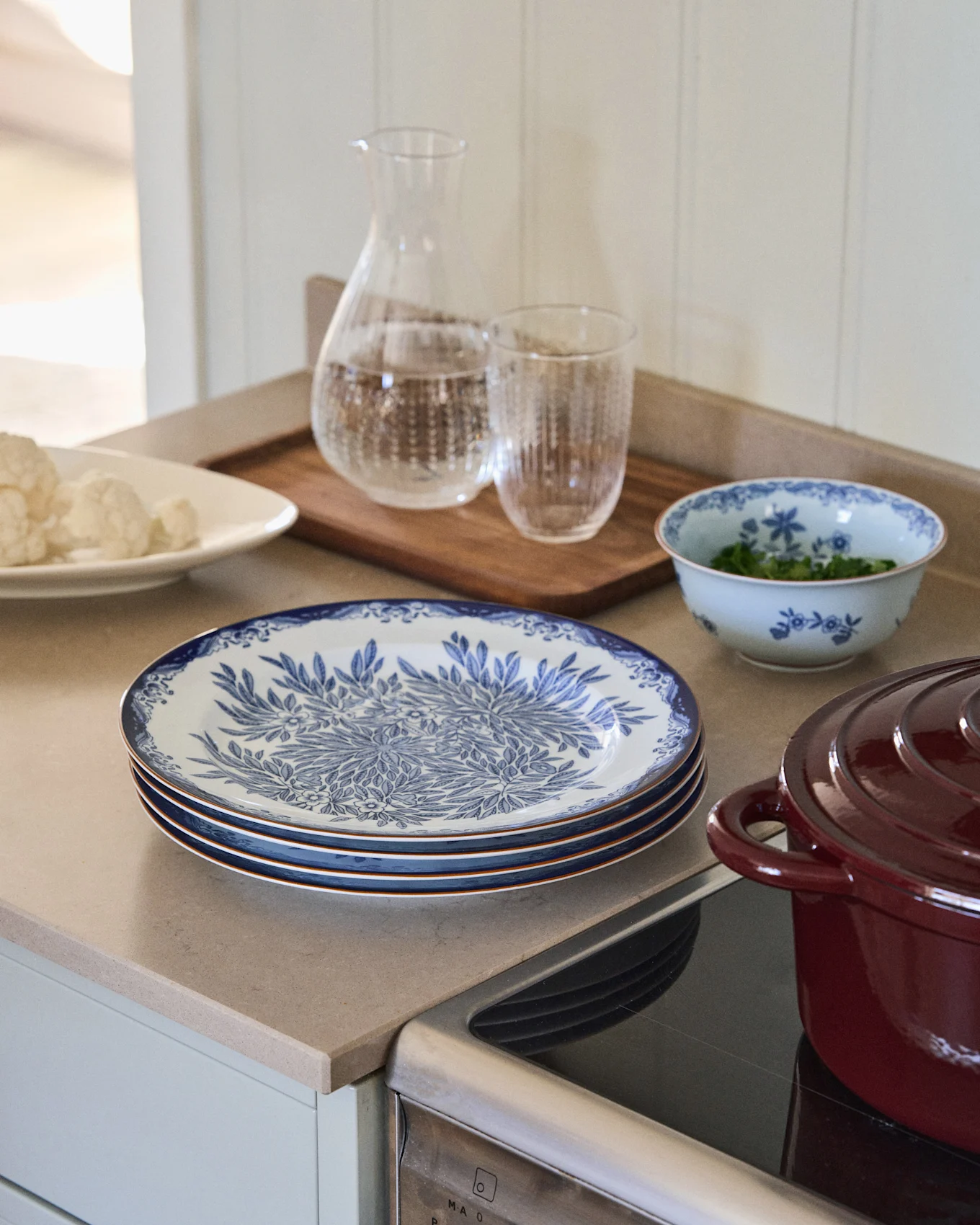 Kitchen counter with blue and white patterned plates, cauliflower, water carafe, a bowl of herbs, and a red pot on a hob.