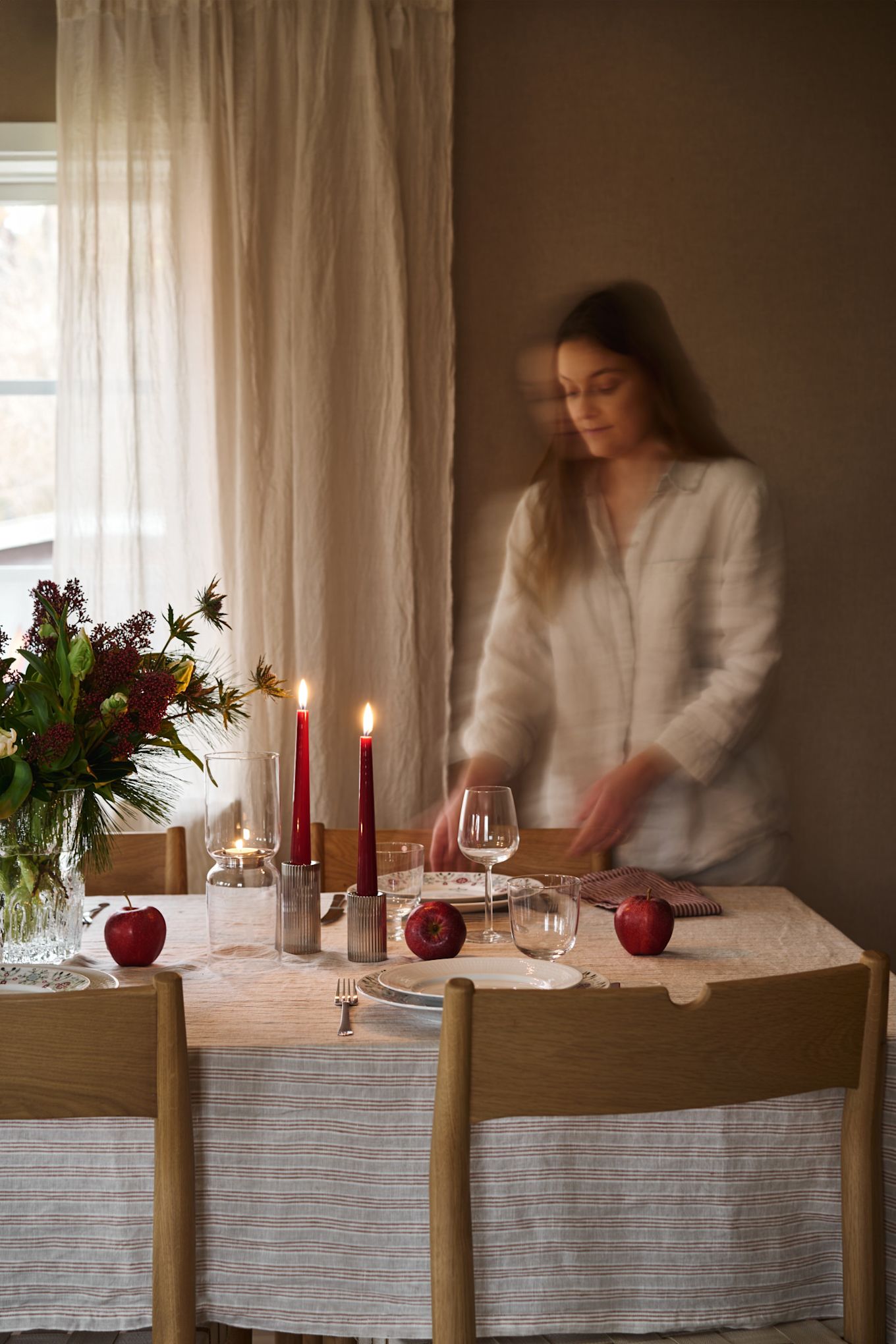 A woman lays a simple Christmas table with red candles, patterned Christmas porcelain and a simple striped tablecloth. 