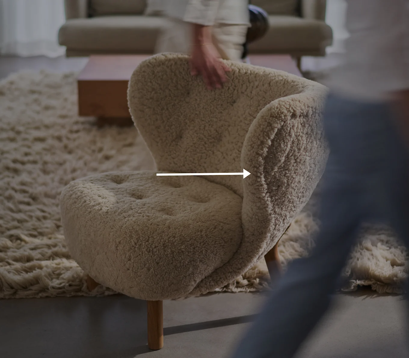 Cream-colored, fluffy armchair with wooden legs in a living room, a blurred hand touching the chair's backrest.