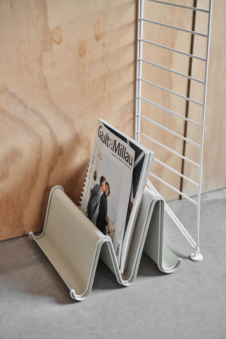 A magazine rack stands on the floor next to a string shelf and a wooden wall. Magazine racks are a great way to organise the living room. 