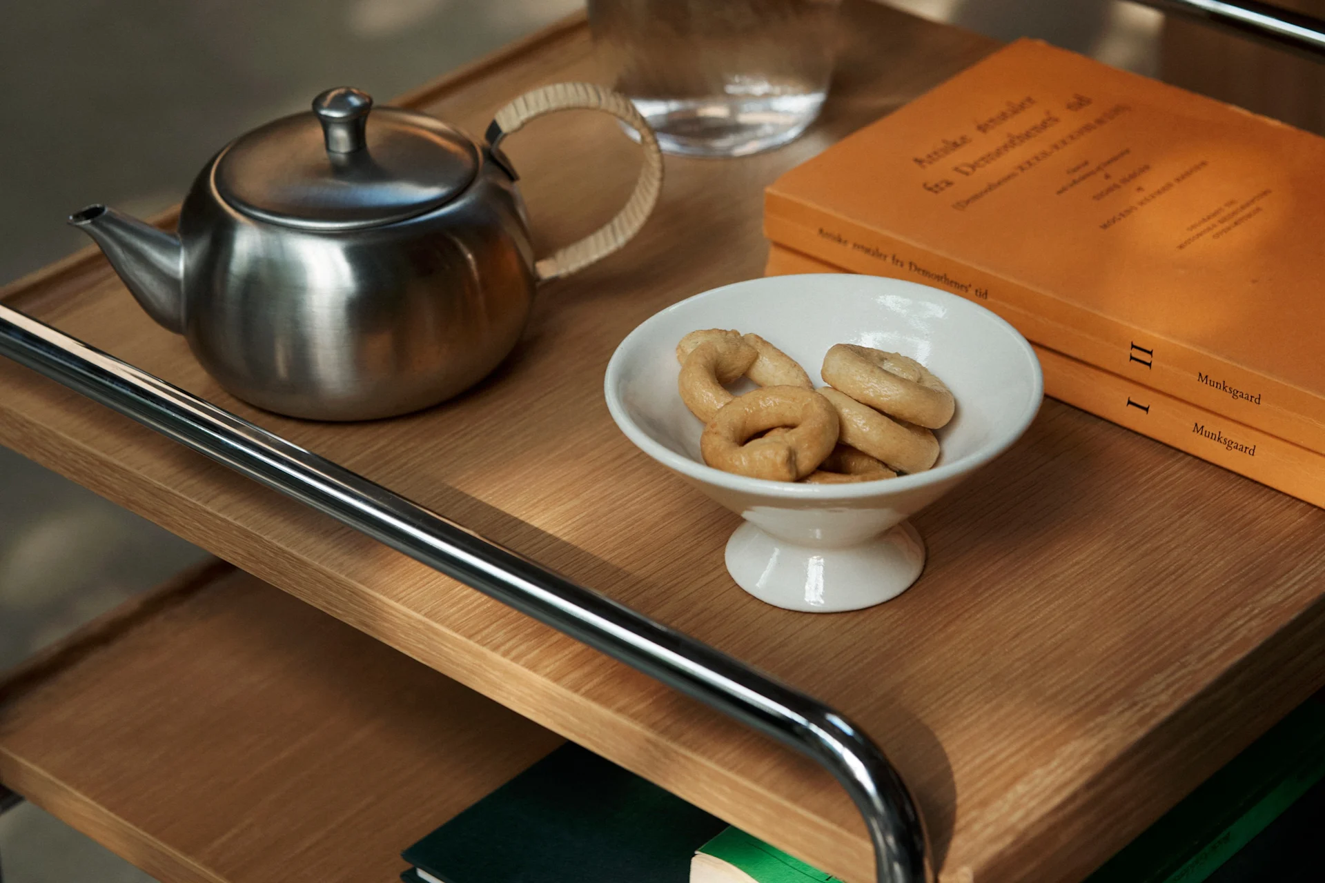 A stainless steel teapot, a white bowl with ring biscuits, and two orange books on a wooden shelf.