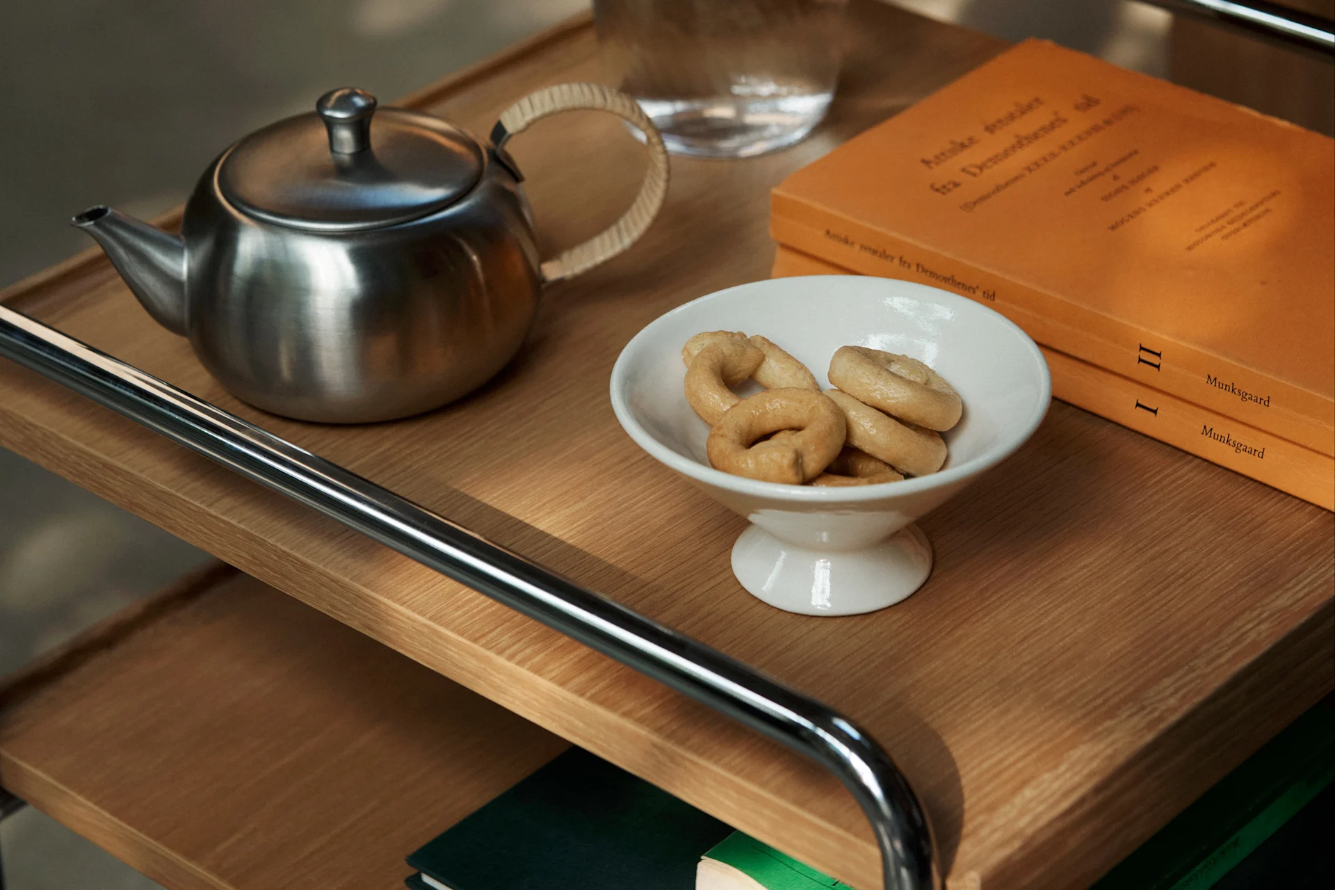 A metal teapot, white bowl of ring cookies, and two orange books on a wooden trolley.
