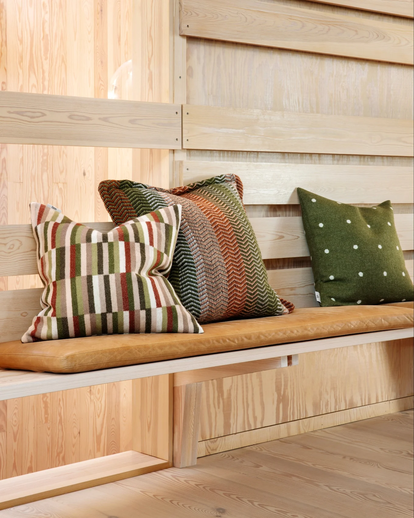 Three throw pillows with different patterns on a light brown leather bench cushion, set against a wooden wall.
