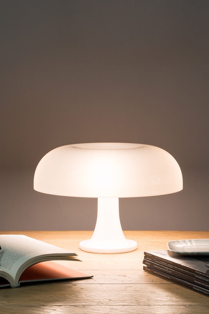 The Nessino lamp illuminated on a desk with an open magazine and a stack of books. 