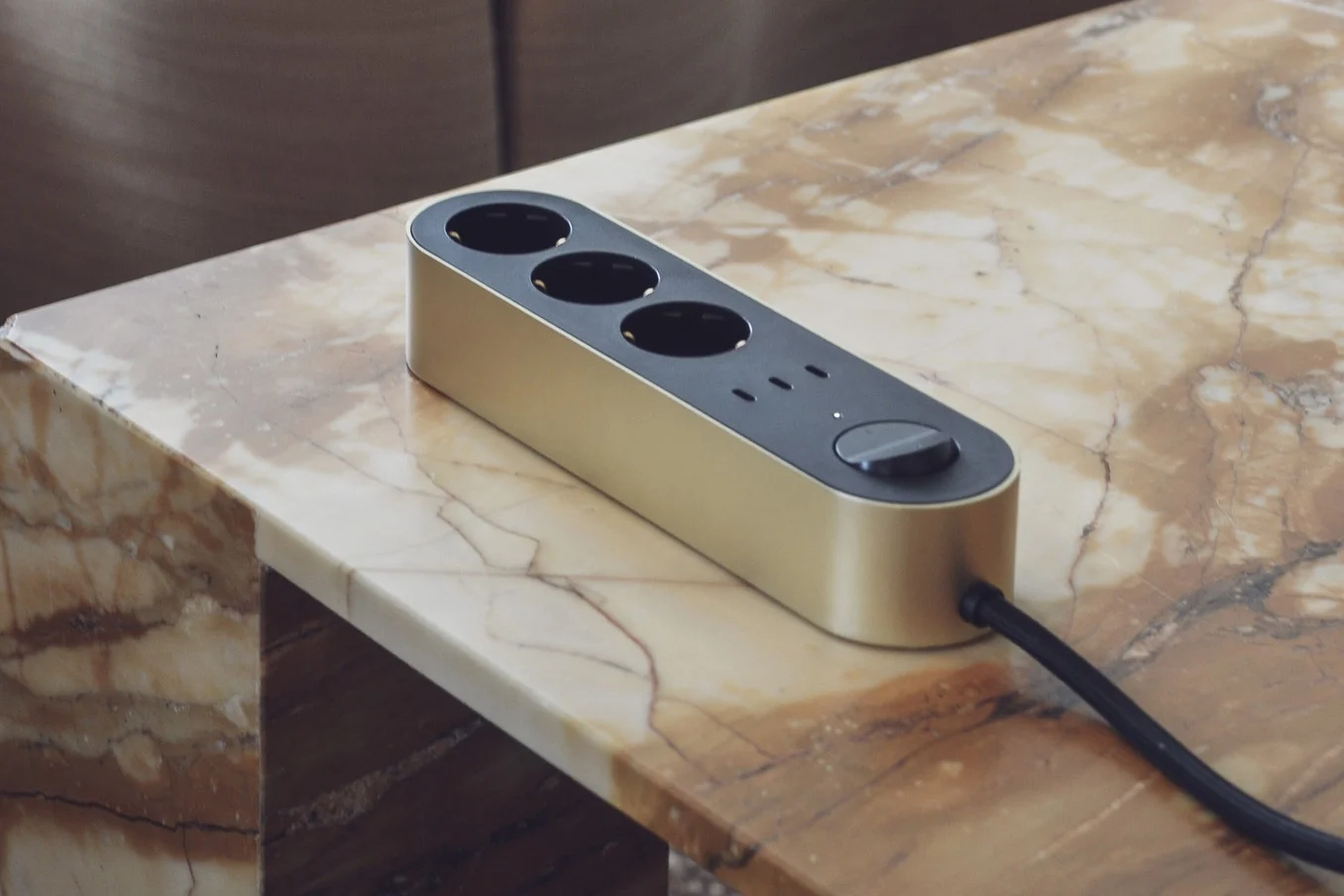 A gold power strip with three black outlets, a control knob, and an indicator light sits on a light brown marble table.
