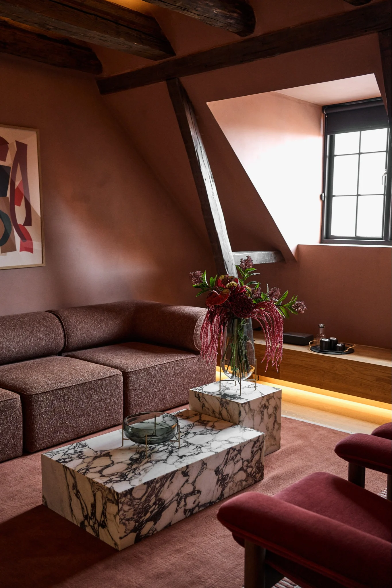 Cozy attic living room with a brown modular sofa, marble coffee tables, dark red armchair, and floral arrangement.