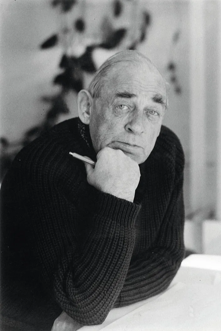 Black and white portrait of Alvar Aalto in a dark sweater, resting his chin on his hand and holding a pen, looking intently at the viewer.
