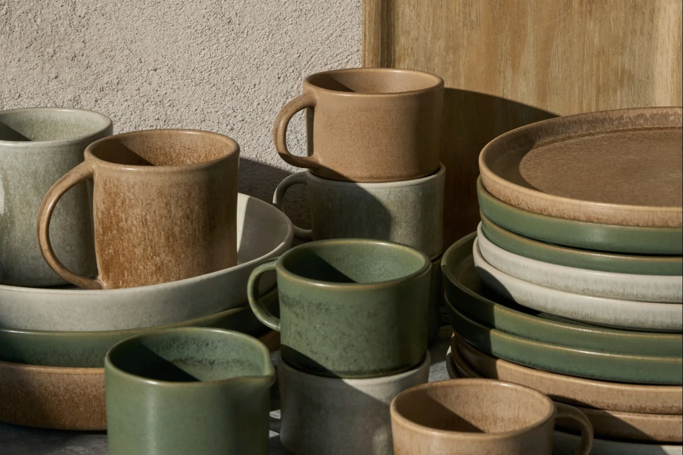 Stacked ceramic mugs, bowls, and plates in beige, light grey, and sage green, with a rustic finish.