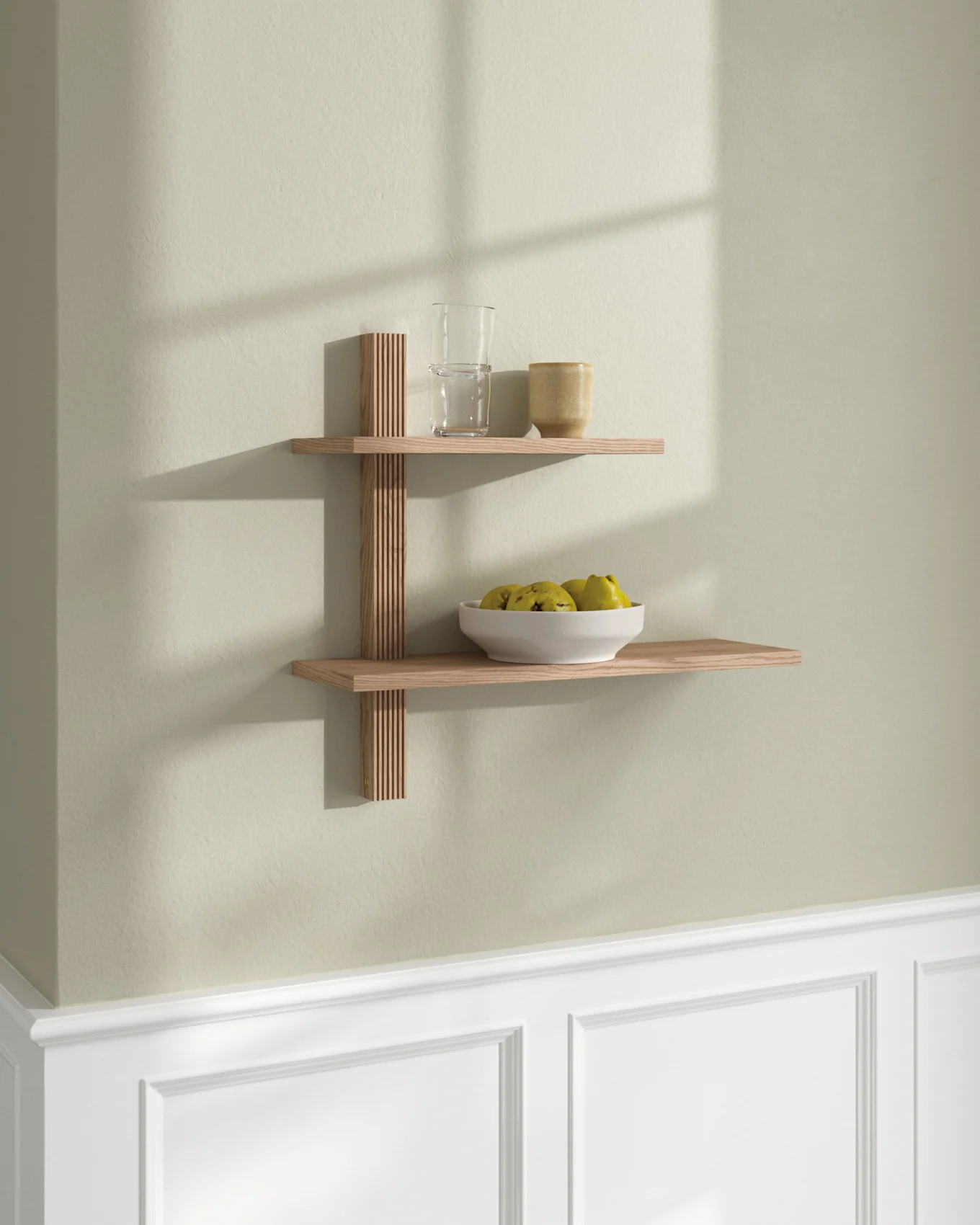 Two light wood floating shelves on a light green wall. Top shelf has a glass and mug; bottom shelf has a white bowl of fruit.