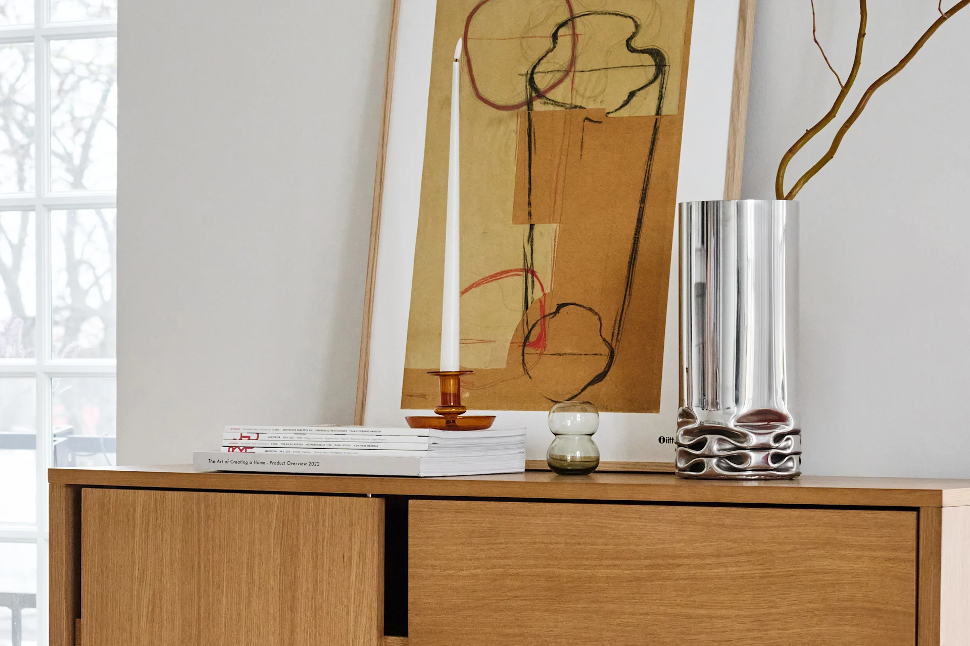 A wooden sideboard holds a framed abstract painting, a white candle in an amber holder, and a silver vase with branches.