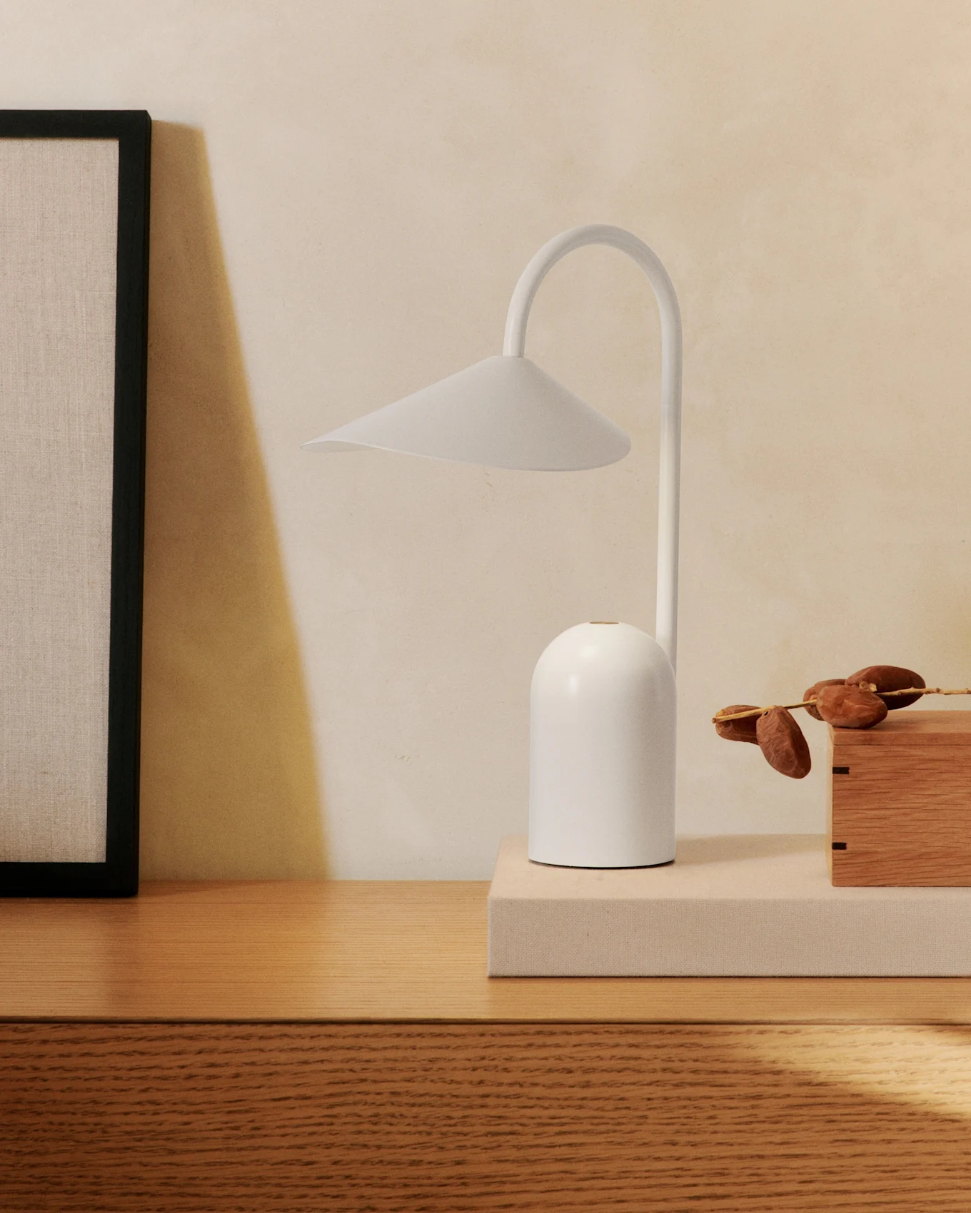 A minimalist white table lamp with a curved arm on a wooden dresser, next to a framed picture and a wooden box with dates.