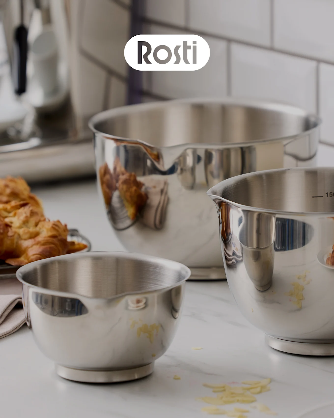Three Rosti bowls on a counter: a beige bowl with a blue one stacked on top, both with lids, and a small yellow bowl filled with dried apricots.