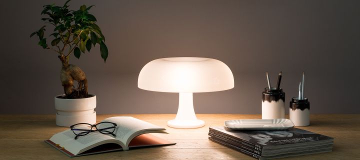 The Nessino mushroom lamp stands illuminated on a desk with pens, books and a plant. 