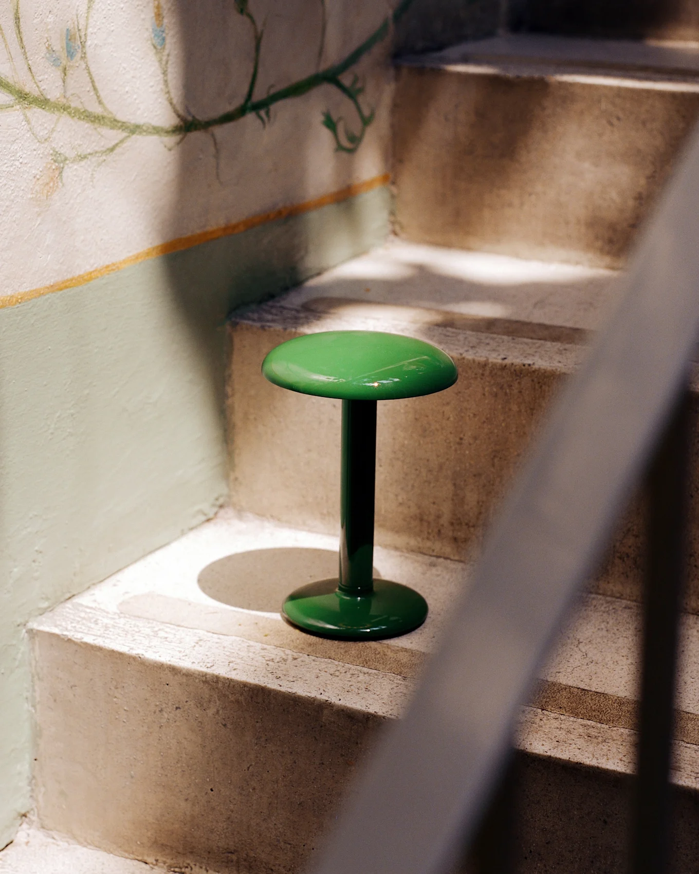 A glossy, bright green lamp with a round top and base stands on a concrete staircase next to a wall with a faded floral mural.