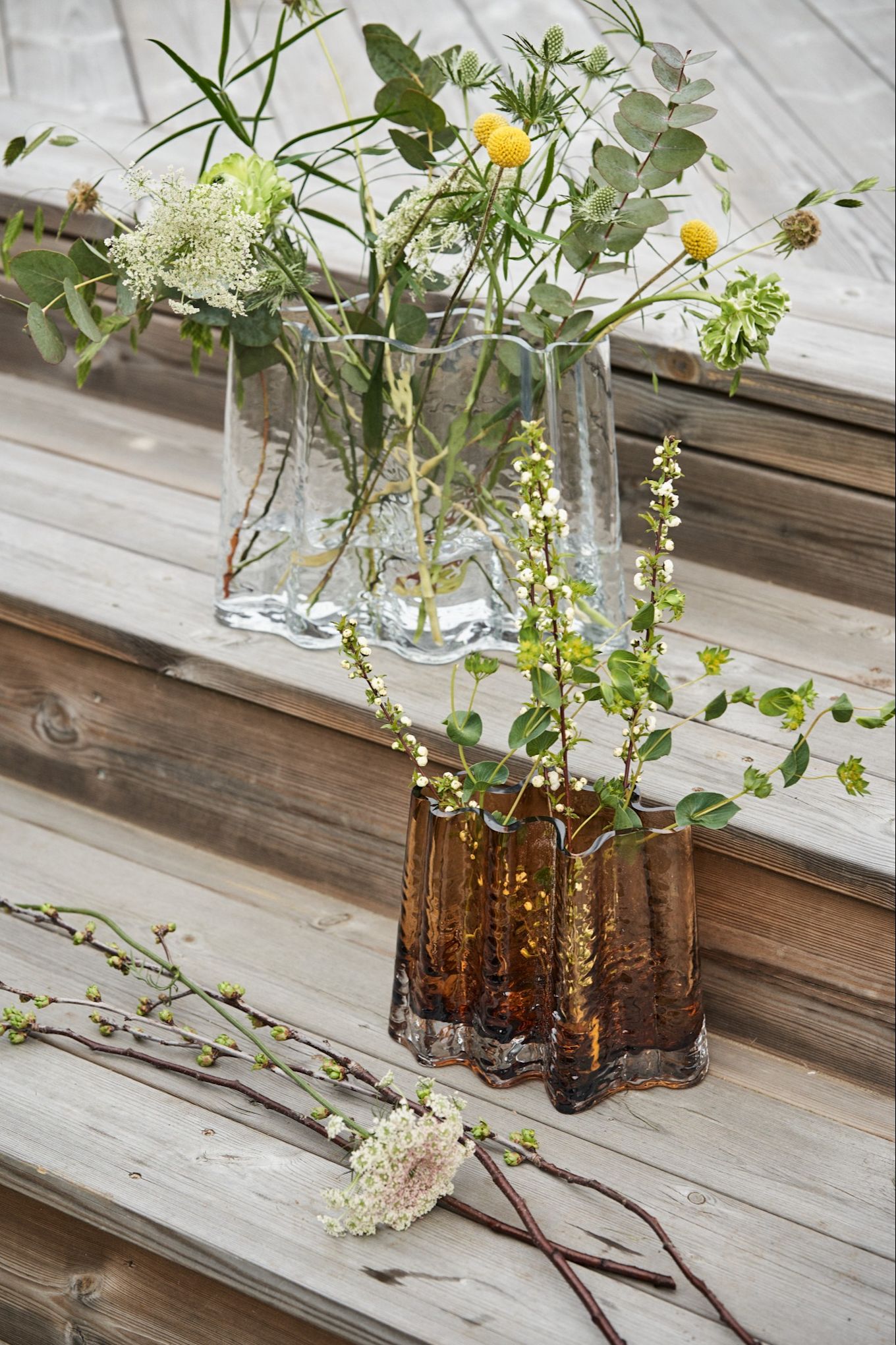 The Gry vase in clear and cognac stand on a wooden deck surrounded by spring flowers. 