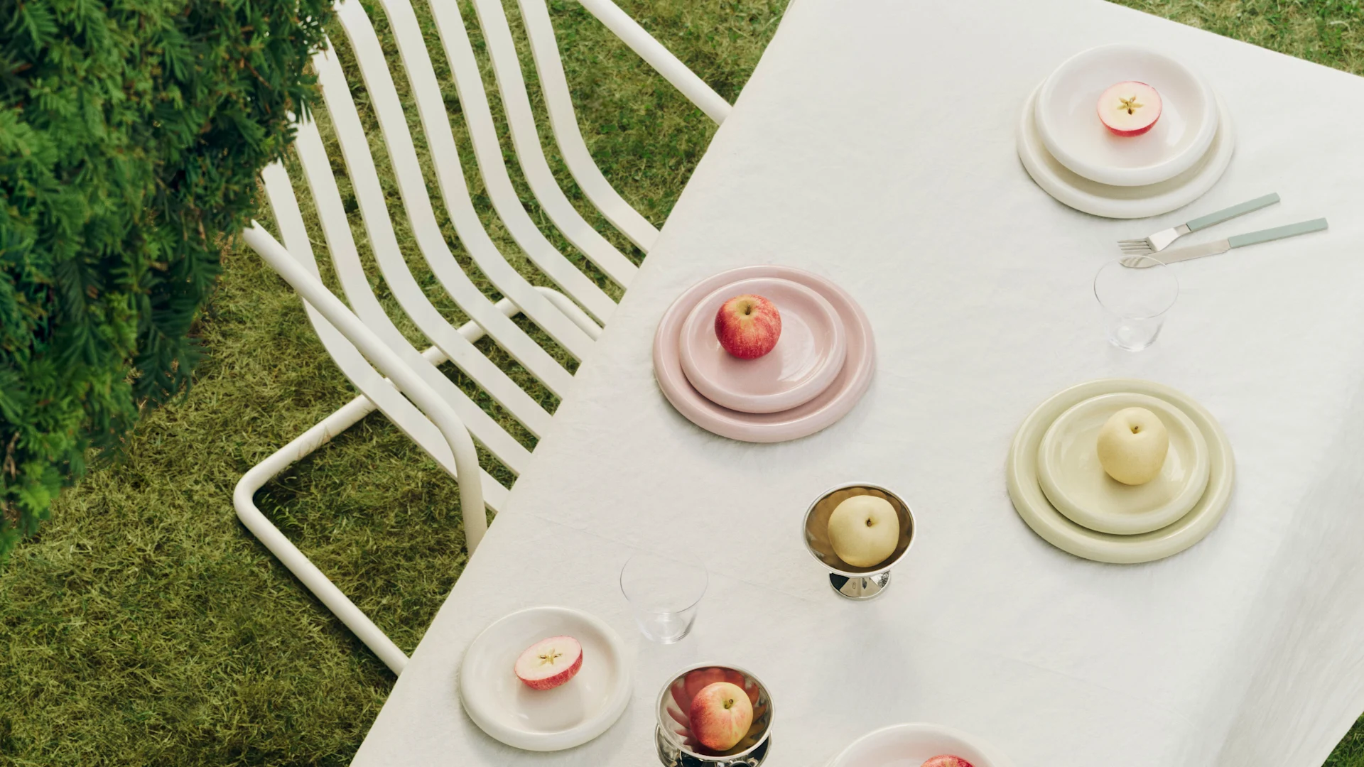 Outdoor table setting with pink, cream, and yellow plates, apples, and cutlery on a white tablecloth.