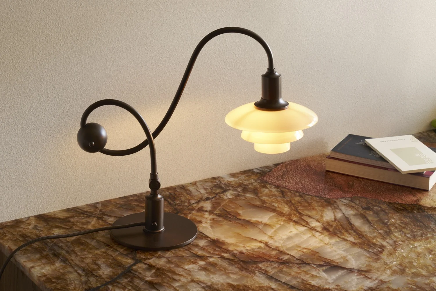 Dark metal table lamp with curved stem and tiered, glowing white shade on a marble-patterned surface with books.