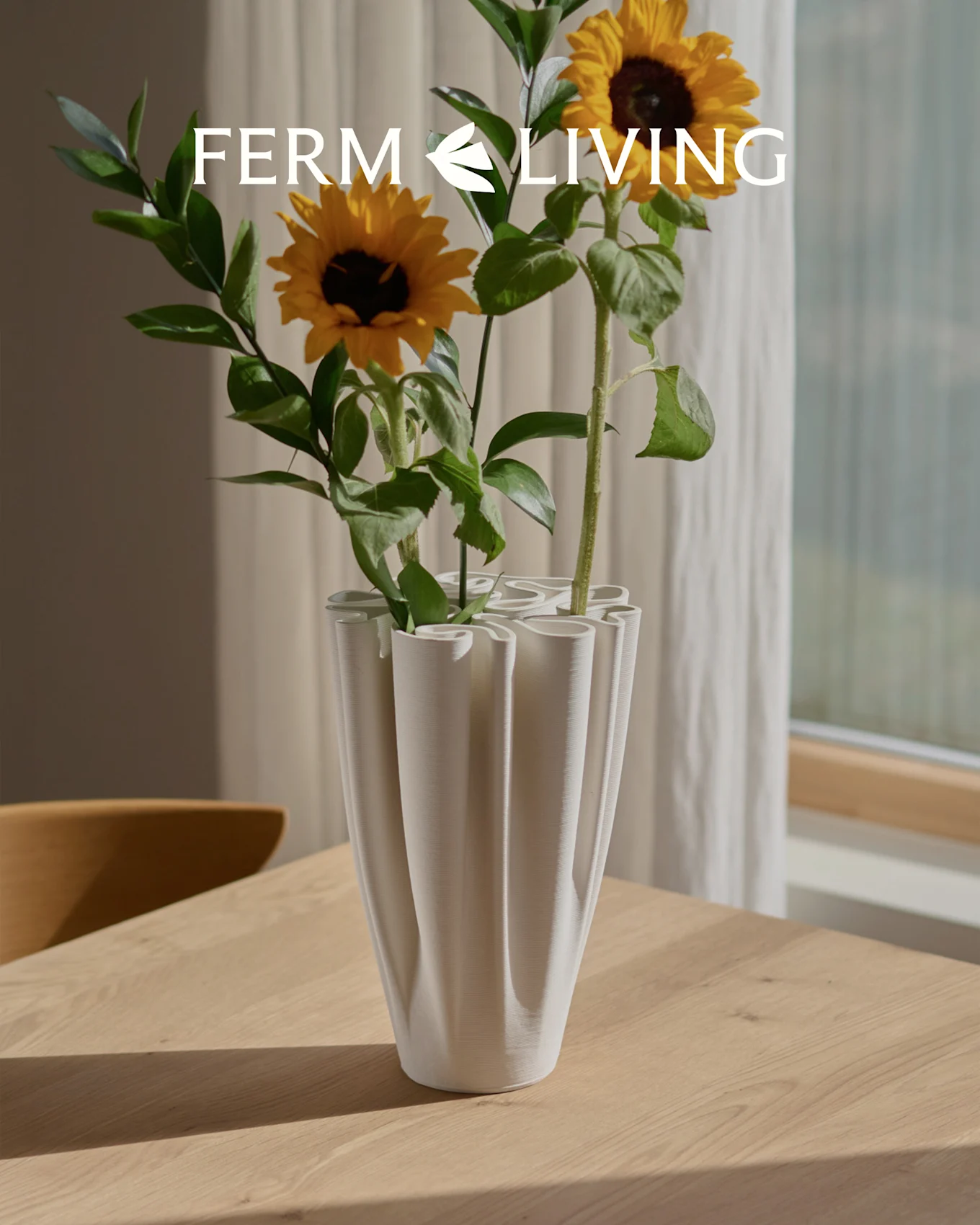 Sunflowers in a white, ribbed wavy vase on a wooden table. Ferm Living logo overlay.