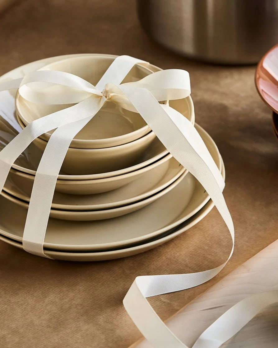 A stack of cream-colored plates and bowls tied with a white ribbon, presented as a gift on brown paper.