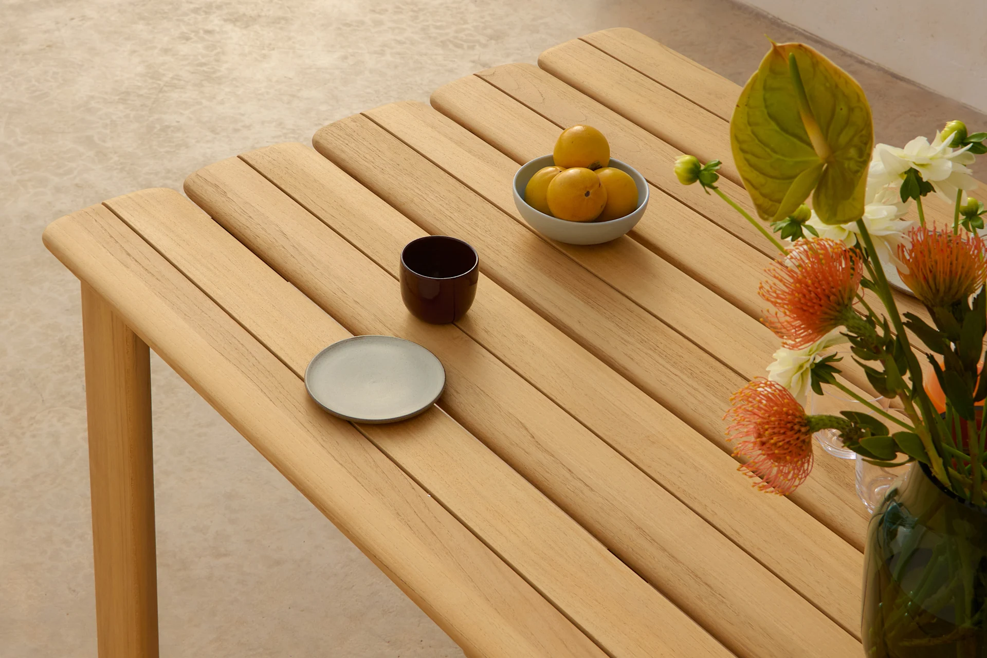A light wooden slatted table with a bowl of yellow fruits, a dark cup, a grey plate, and a vase of orange and white flowers.