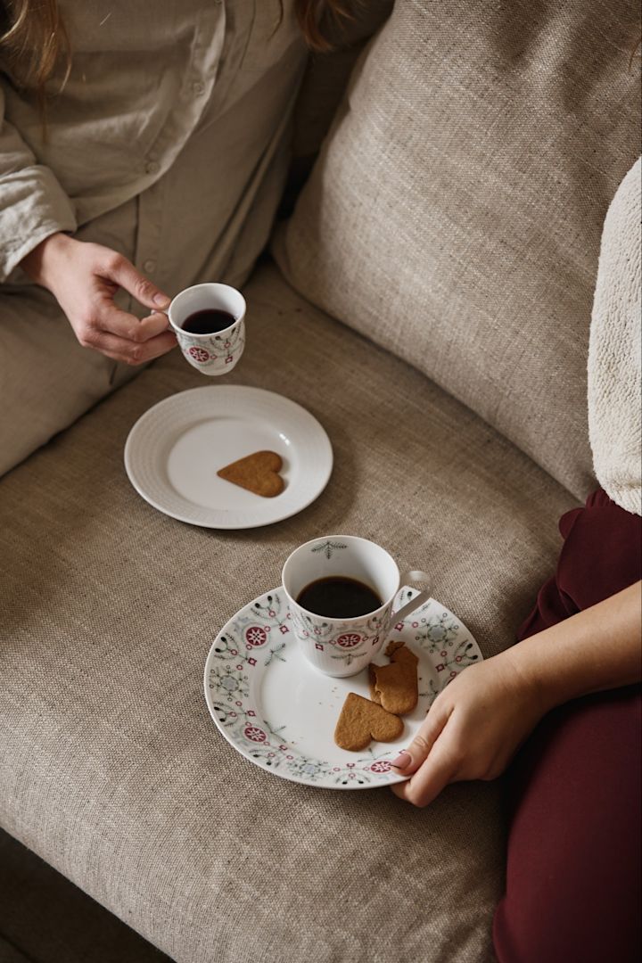 A couple of people sit on a sofa with the Swedish Grace Winter mug collection. Here you see both a regular mug and a glögg mug as well as plates with ginger bread biscuits. 