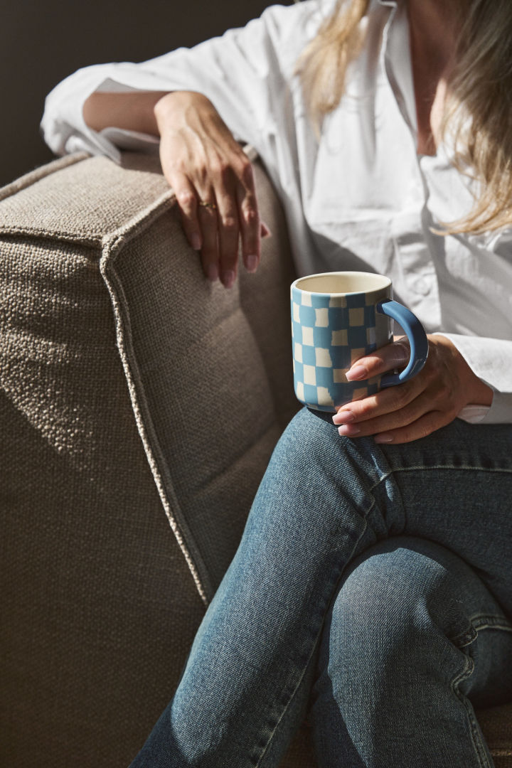 Checks and stripes are set to be a big trend in our interiors in autumn 2024. Here you see a woman sat on a chair in the sunshine holding a blue checked mug from Byon. 