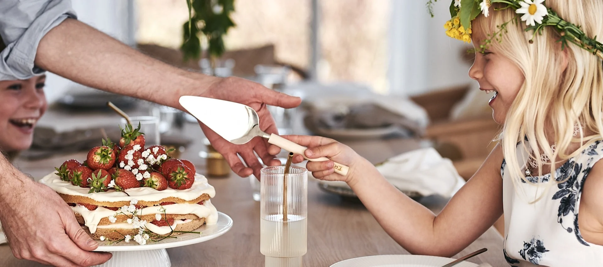 Discover Swedish midsummer party ideas, a child cuts into a traditional strawberry cake while wearing a midsummer flower crown.