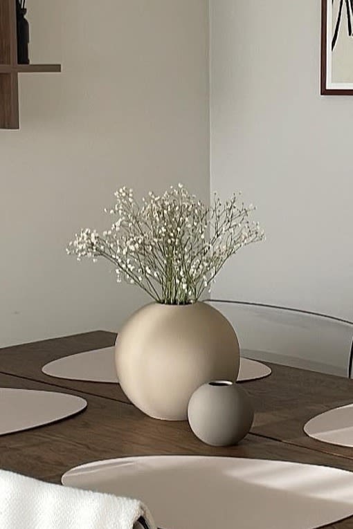 The Ball vase in sand and grey, in two different sizes stands on a dining room table in a neutral-coloured dining room. 