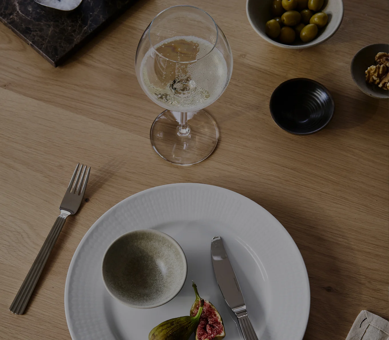 A glass of champagne, white plate with a bowl and figs, fork, knife, olives, and walnuts on a wooden table.
