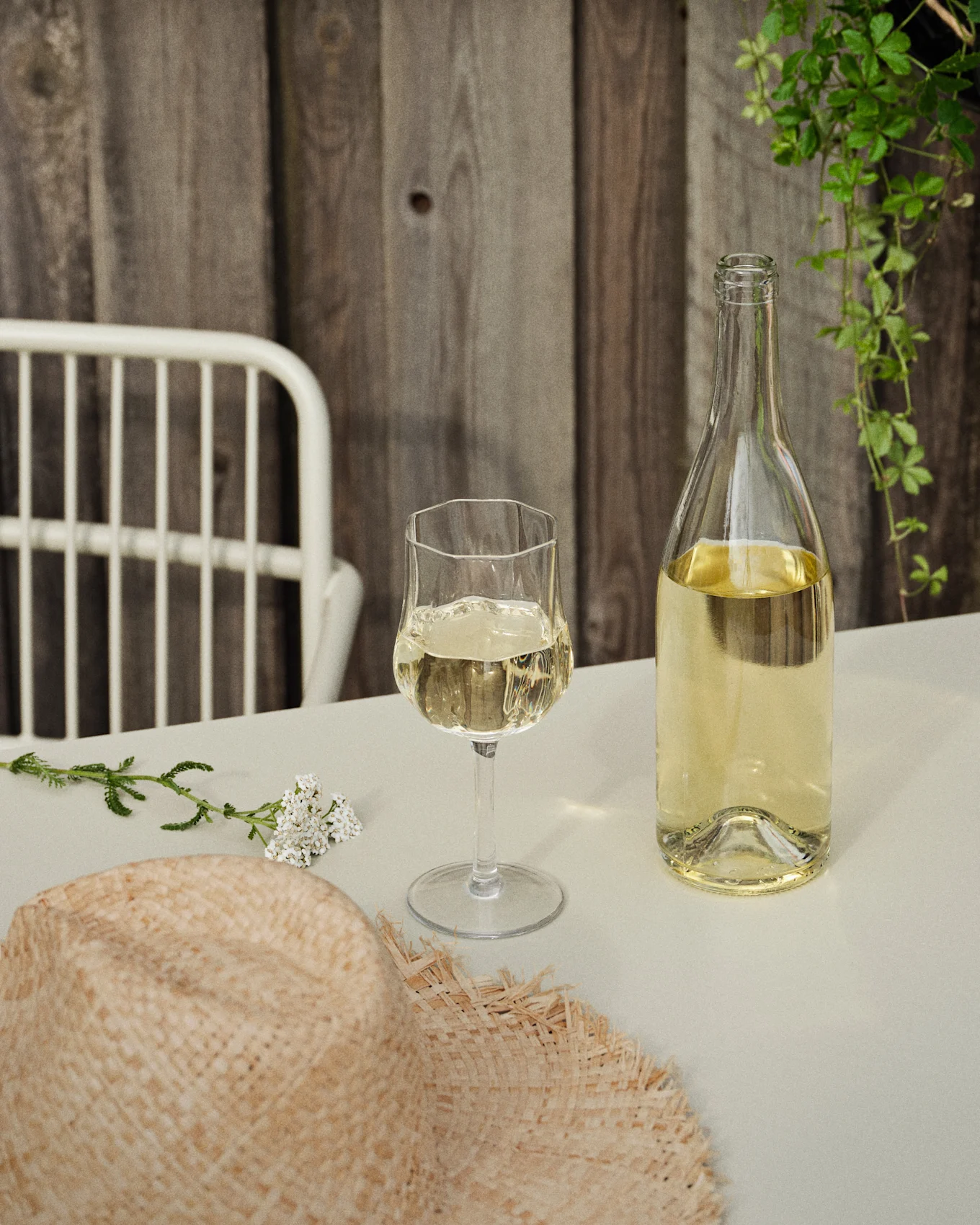 A bottle and glass of white wine on a table with a straw hat and white flowers, against a wooden wall and green vines.