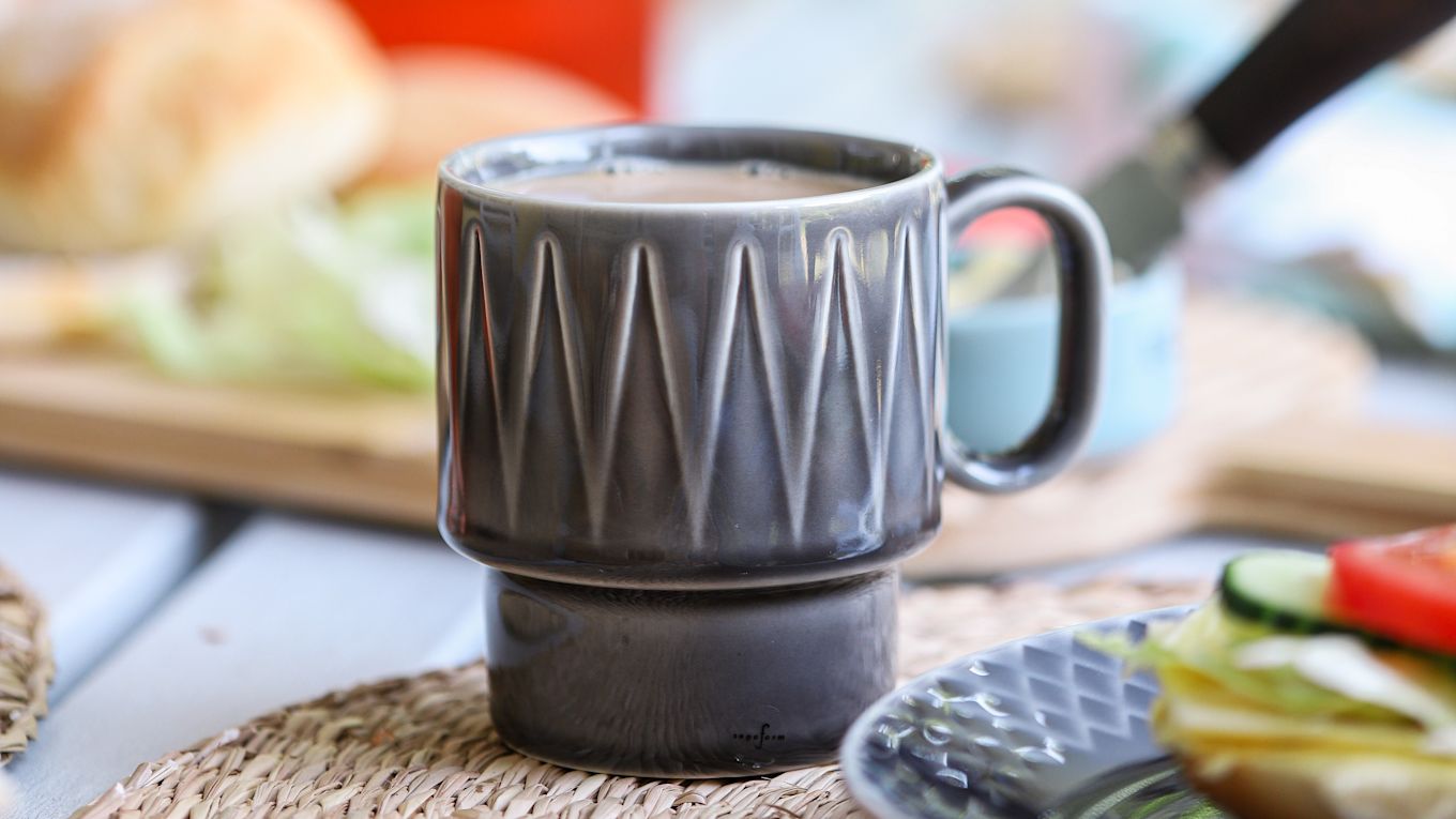 Image showing the Coffee Moore coffee mug in grey from Sagaform, filled with coffee, next to a plate with freshly baked bread.