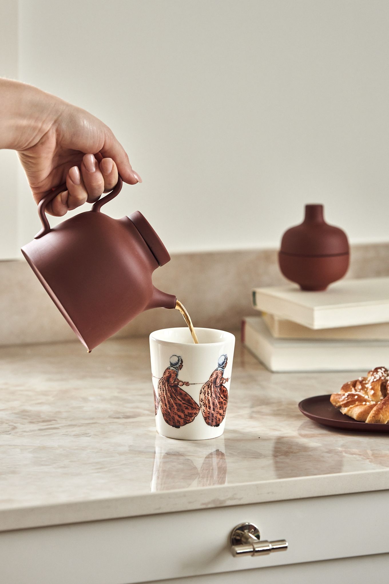 Mug with the Swedish author Elsa Beskow motif Aunt Brown on a kitchen counter together with the pitcher Sand in terracotta-red.