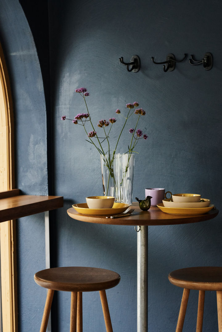 Hier zie je een retro tafeldecoratie in geel en paars in een donkerblauwe kamer met een heldere vaas en bijpassende paarse bloemen. 