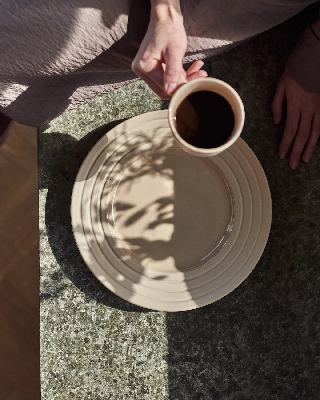 Overhead view: hands, one holding a beige mug with black coffee over a beige plate on a dark stone table, with leaf shadows.
