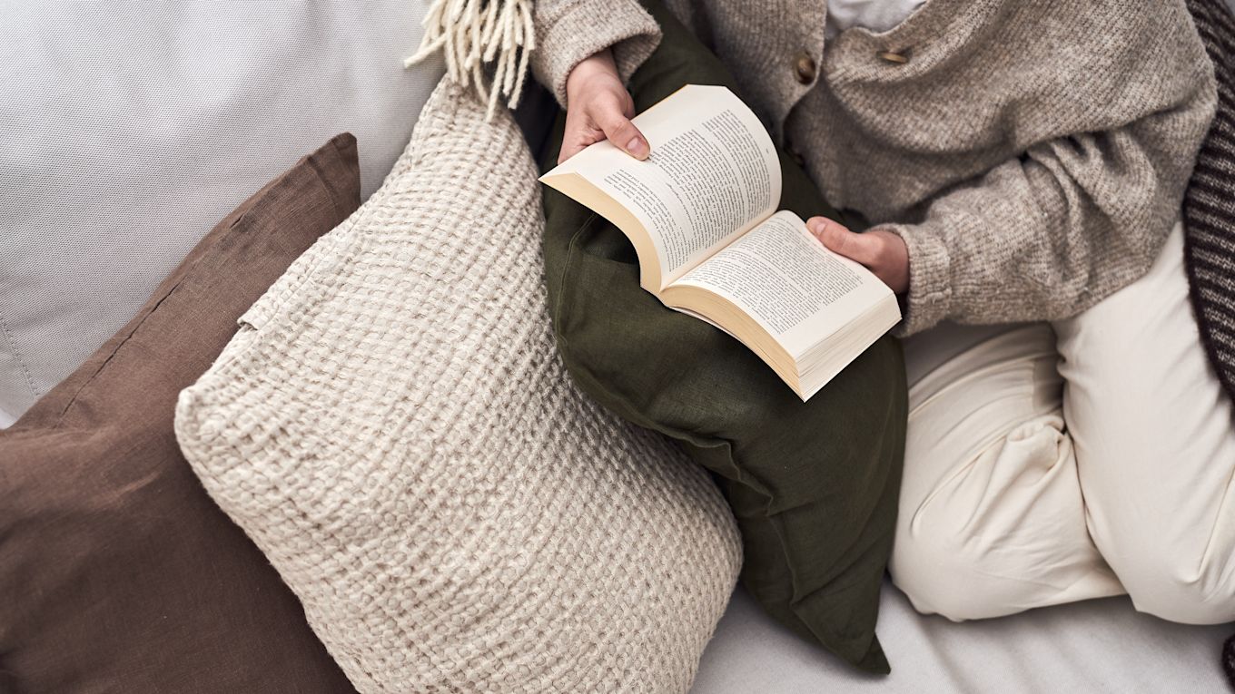 A woman curls up on a small outdoor sofa on a balcony with a book. 