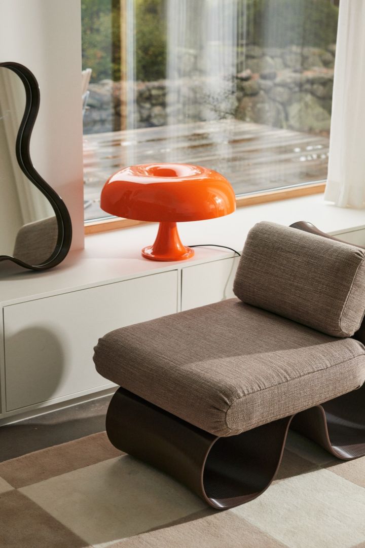 The Nessino lamp in orange with it's doughnut shaped lamp shade stands in a window with a curved edge mirror and a matching lounge chair. 