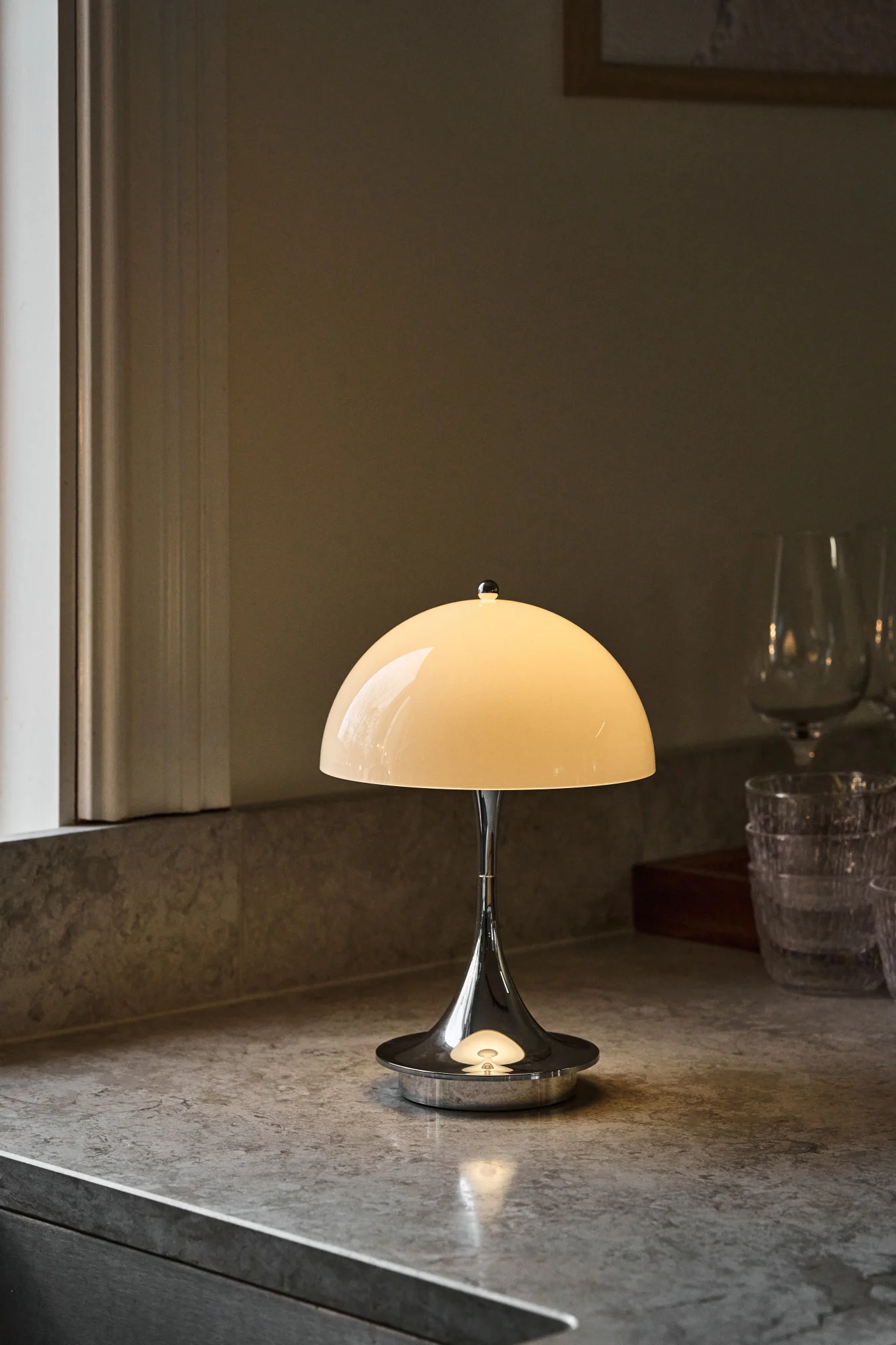 Polished chrome table lamp with a glowing cream dome shade on a speckled gray countertop, next to a window and blurred glasses.