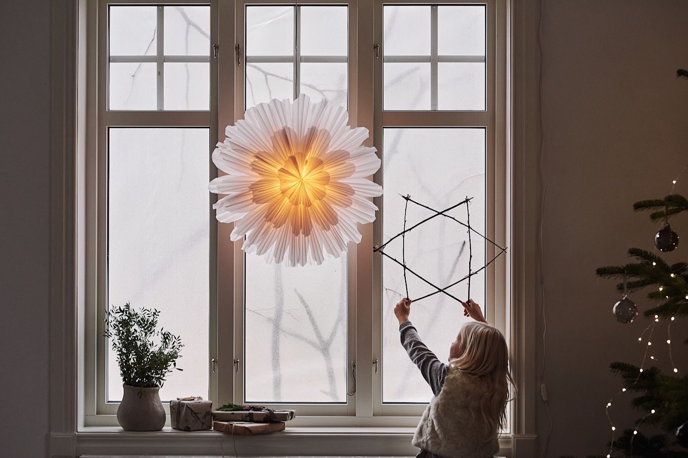 Snöblomma Christmas star from Watt & Veke in a window with a child holding a star made from of sticks.