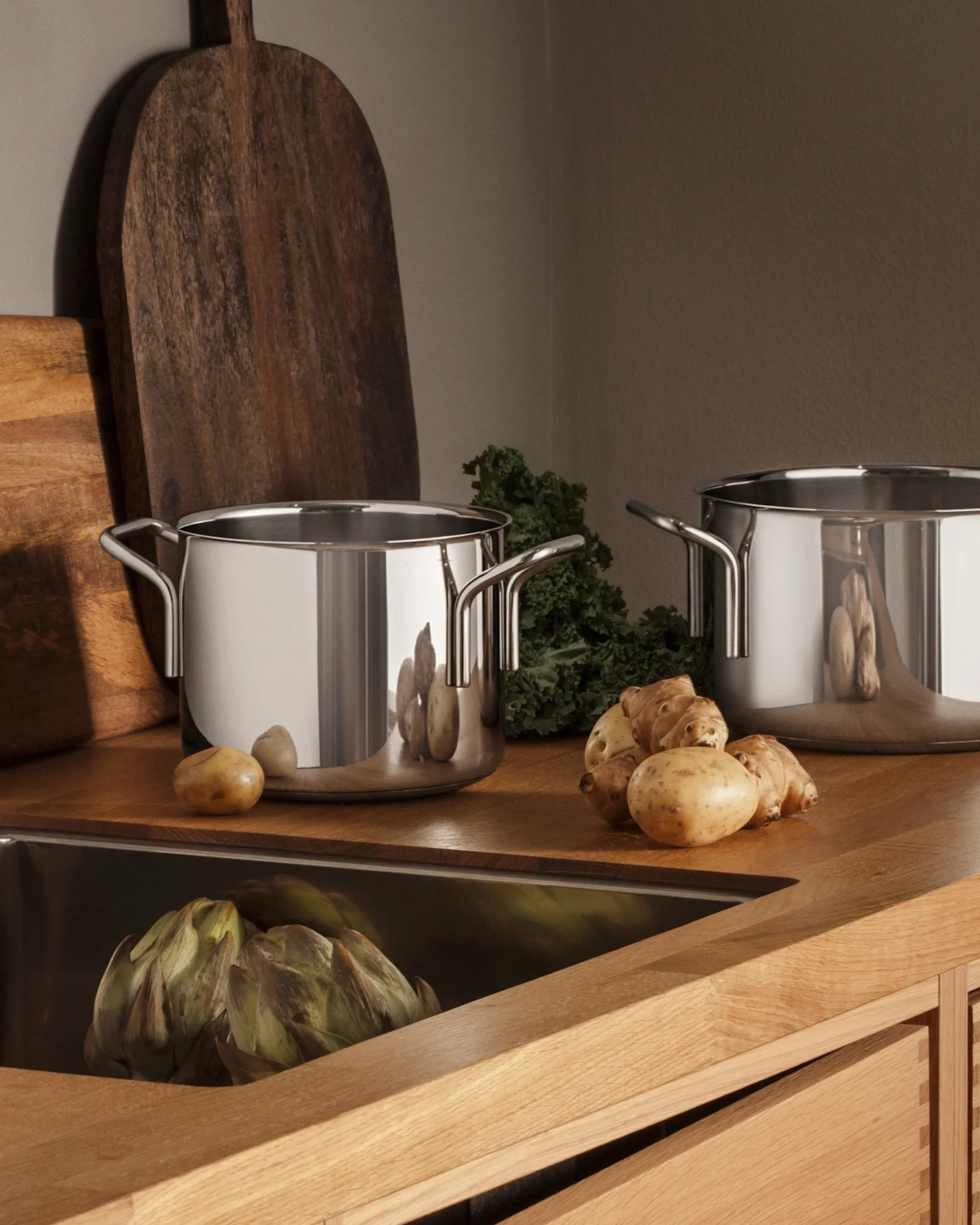 Modern kitchen with wooden counter, stainless steel sink, faucet, three pots, and fresh produce like potatoes and artichokes.