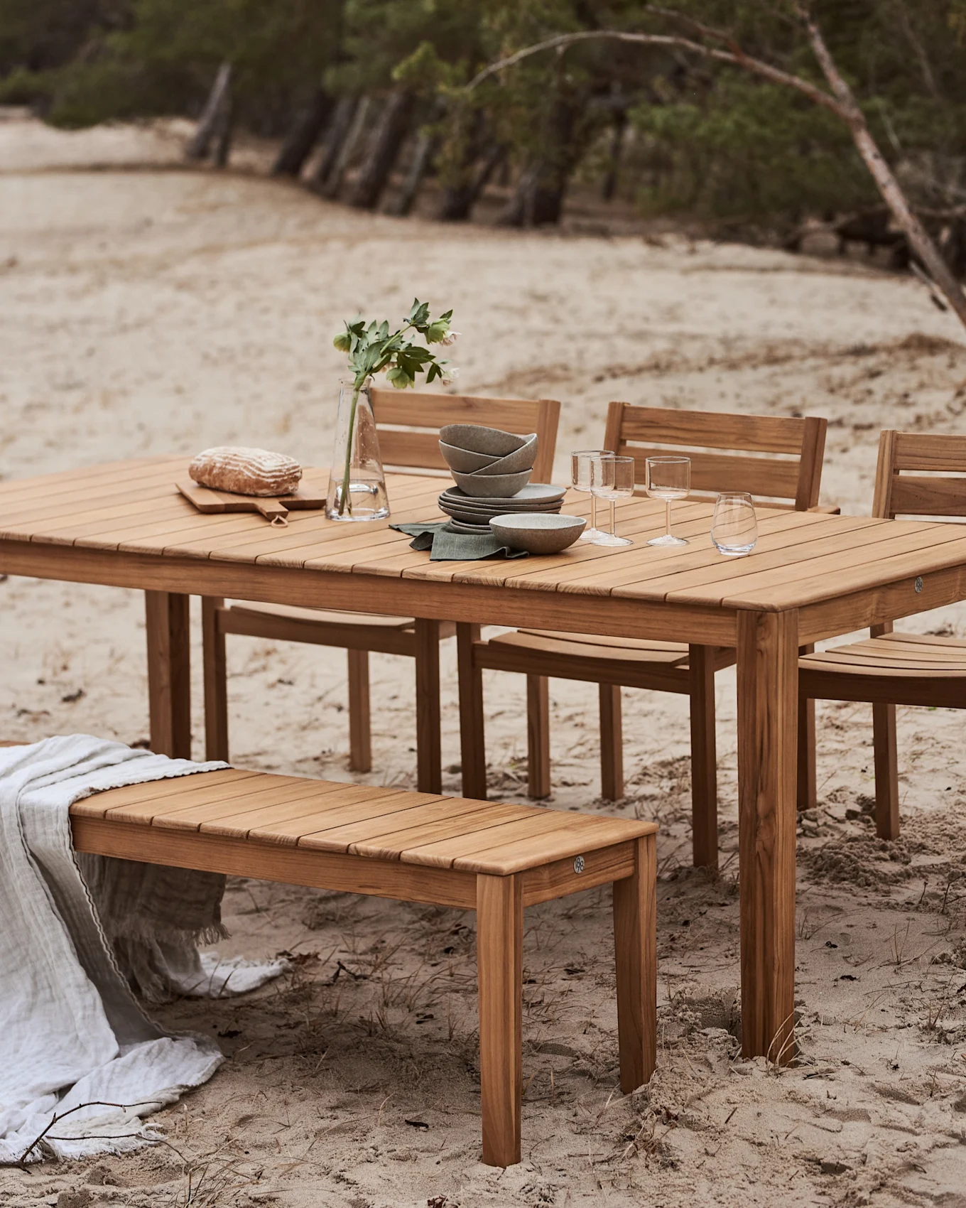 Outdoor wooden dining set with a table and bench on a sandy beach. The table is set with dishes, glasses, bread, and a vase with flowers.