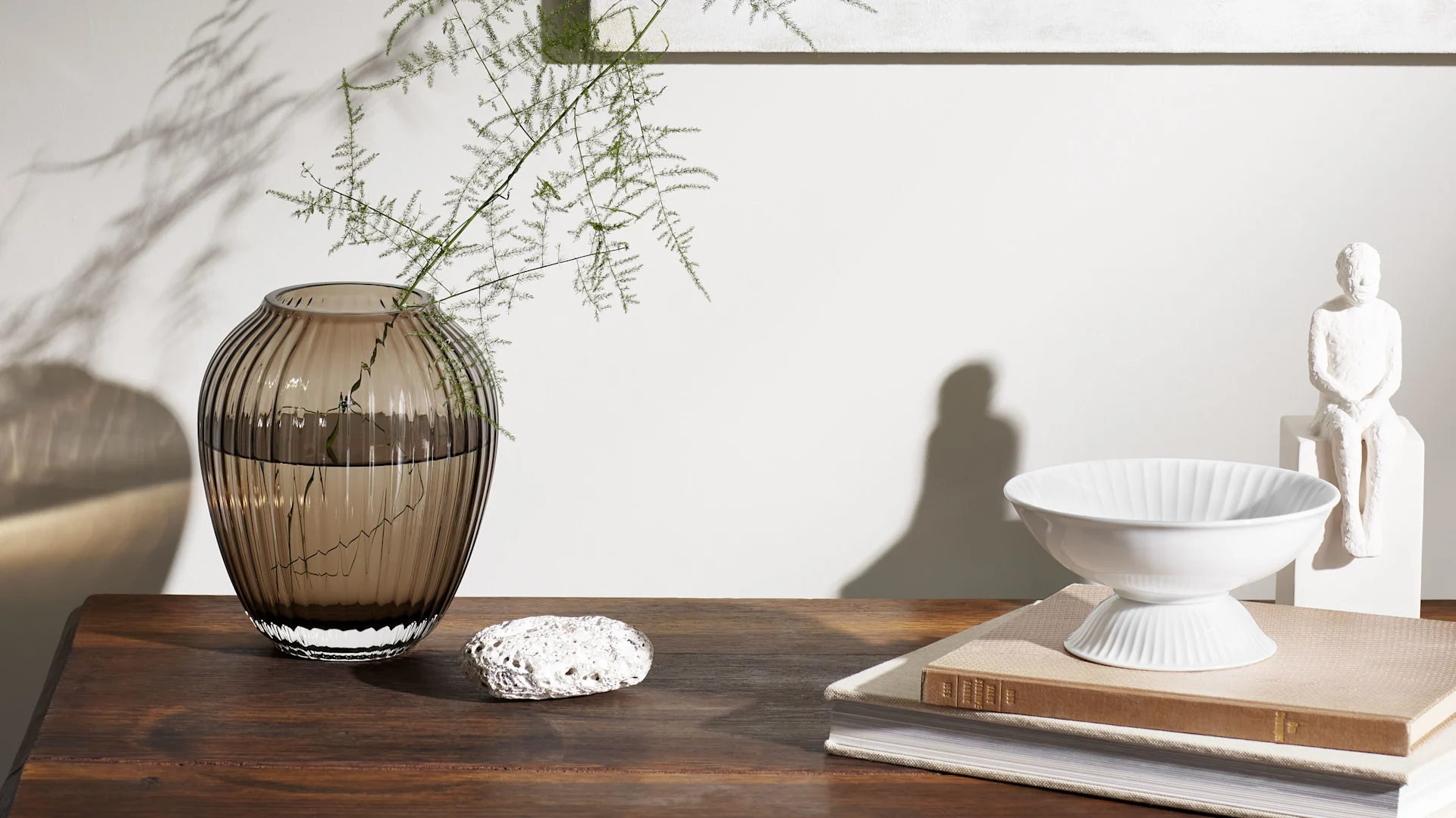 Still life: smoky ribbed vase with greenery, white textured rock, fluted bowl, and abstract sculpture on a wooden table.