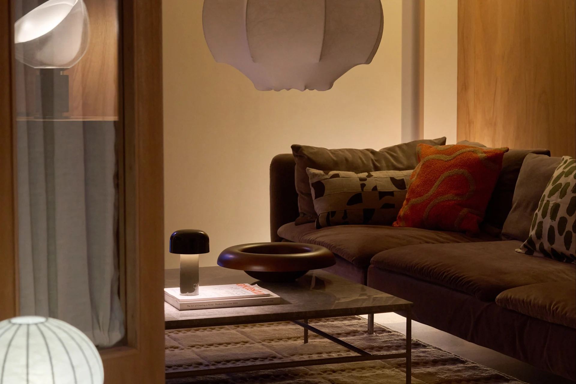 A cozy living room with a brown sofa, a large white pendant lamp, a floor lamp, and a coffee table with a mushroom-shaped lamp and a decorative bowl.