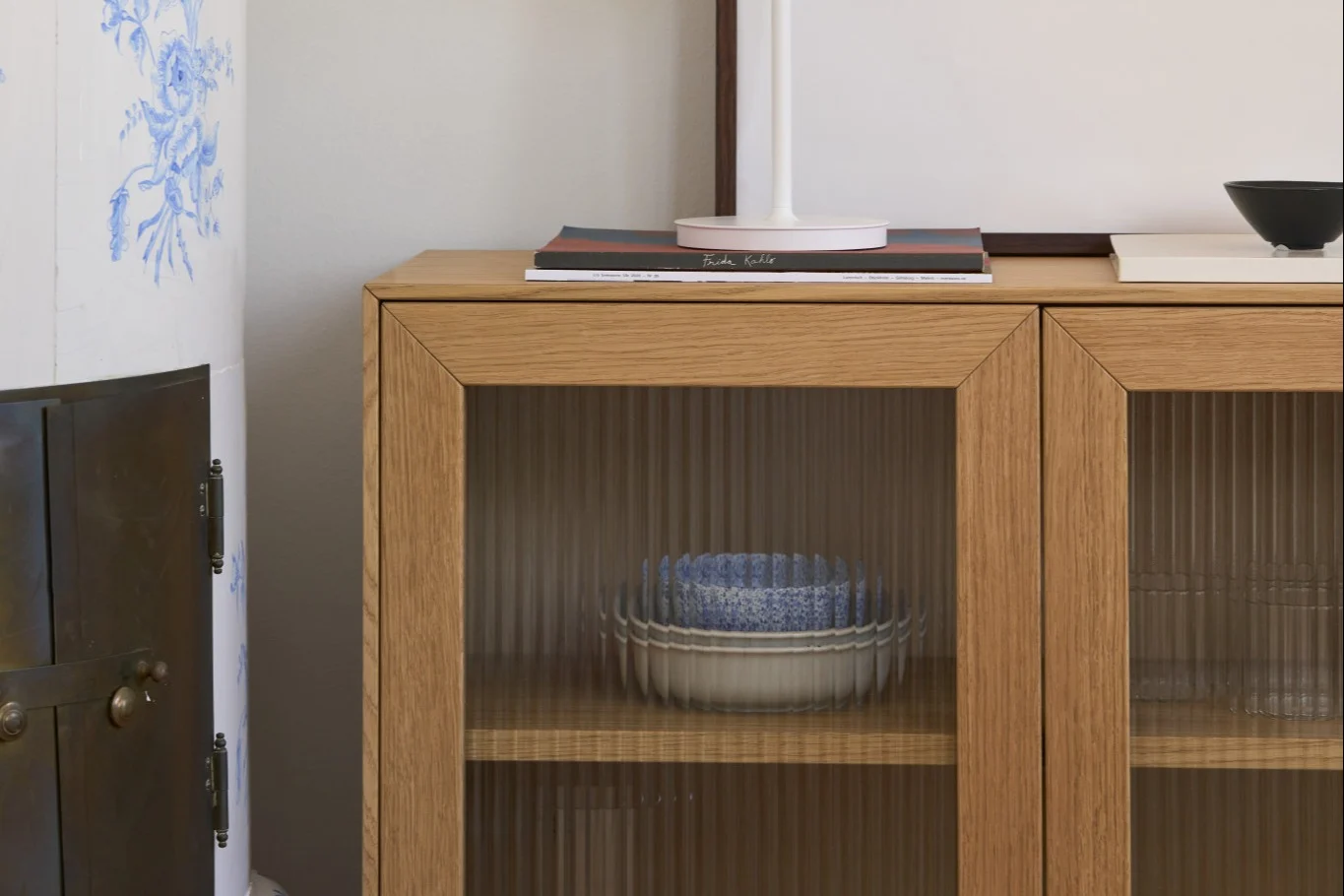 Wooden cabinet with fluted glass doors, featuring stacked blue and white bowls inside, next to a tiled stove.