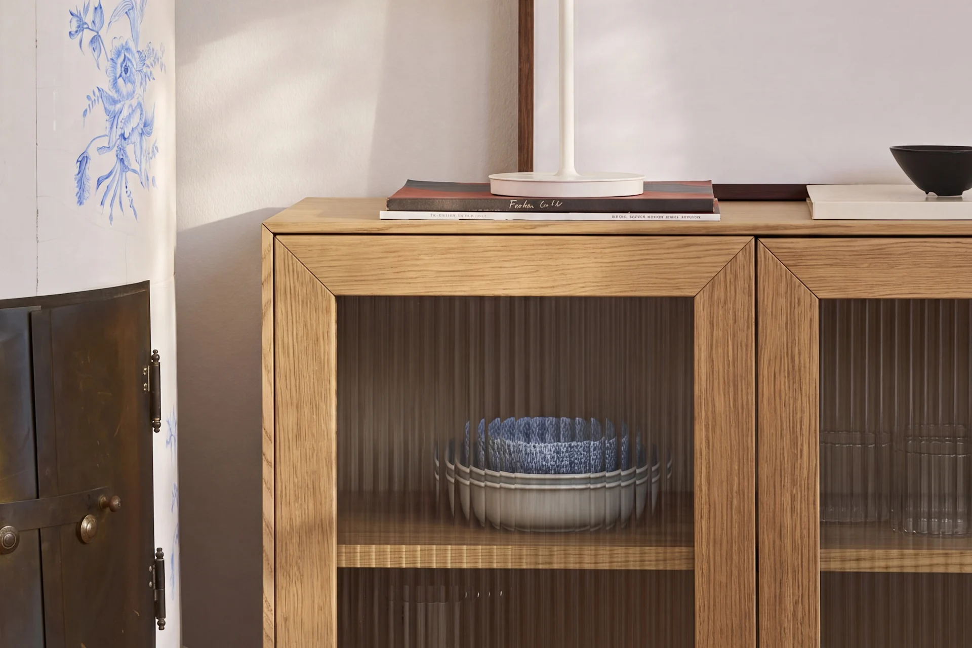 A light wood cabinet with fluted glass doors holds blue and white bowls, next to a ceramic stove with blue floral patterns.